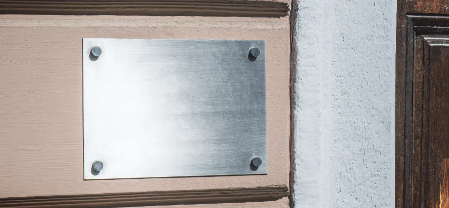 Brushed stainless steel blank sign mounted with four standoff screws on a beige wall beside a wooden door.