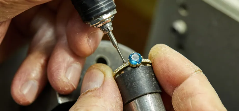 Jeweler using a hand tool to precisely set a blue gemstone into a gold ring with diamond accents.