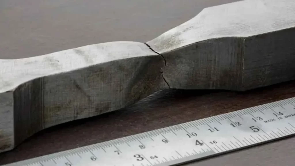 The image shows a broken test specimen with a scale placed next to the fractured part, visually representing material failure.