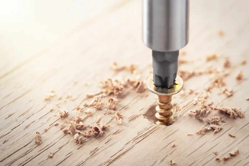 A drill bit inserting a screw into a wooden board with wood shavings scattered around the surface