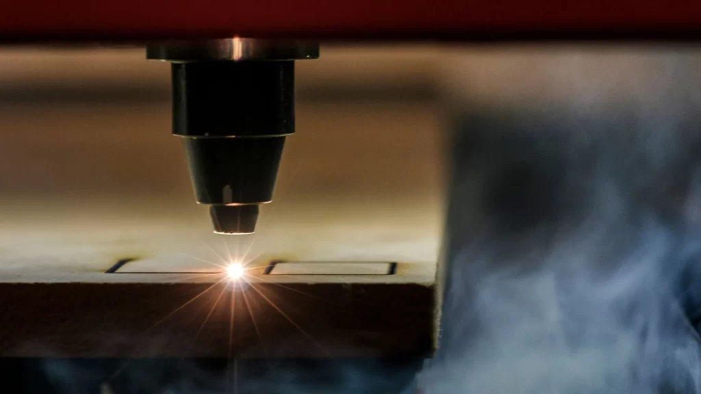 Industrial CO2 laser cutting head in operation, producing bright sparks while precisely cutting through a flat wood workpiece surface.