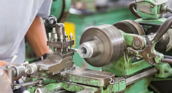 The operator's hands are adjusting the manual lathe machine, working on a cylindrical metal workpiece.